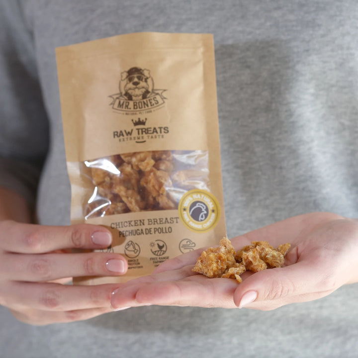 Persona mostrando el snack natural Raw Treats Pechuga de Pollo para perro y gato, con la bolsa de Mr. Bones y los trozos de pollo deshidratado en la mano.